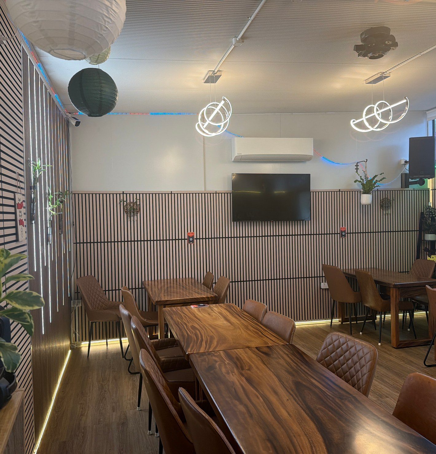 The private room at Windsor Ave: two long live-edge timber tables set with tan leather chairs, paper lanterns overhead and a wall-mounted screen